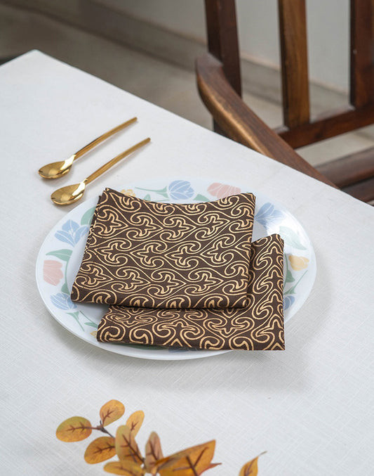Brown patterned napkin on a white plate with gold spoons, placed on a tablecloth.